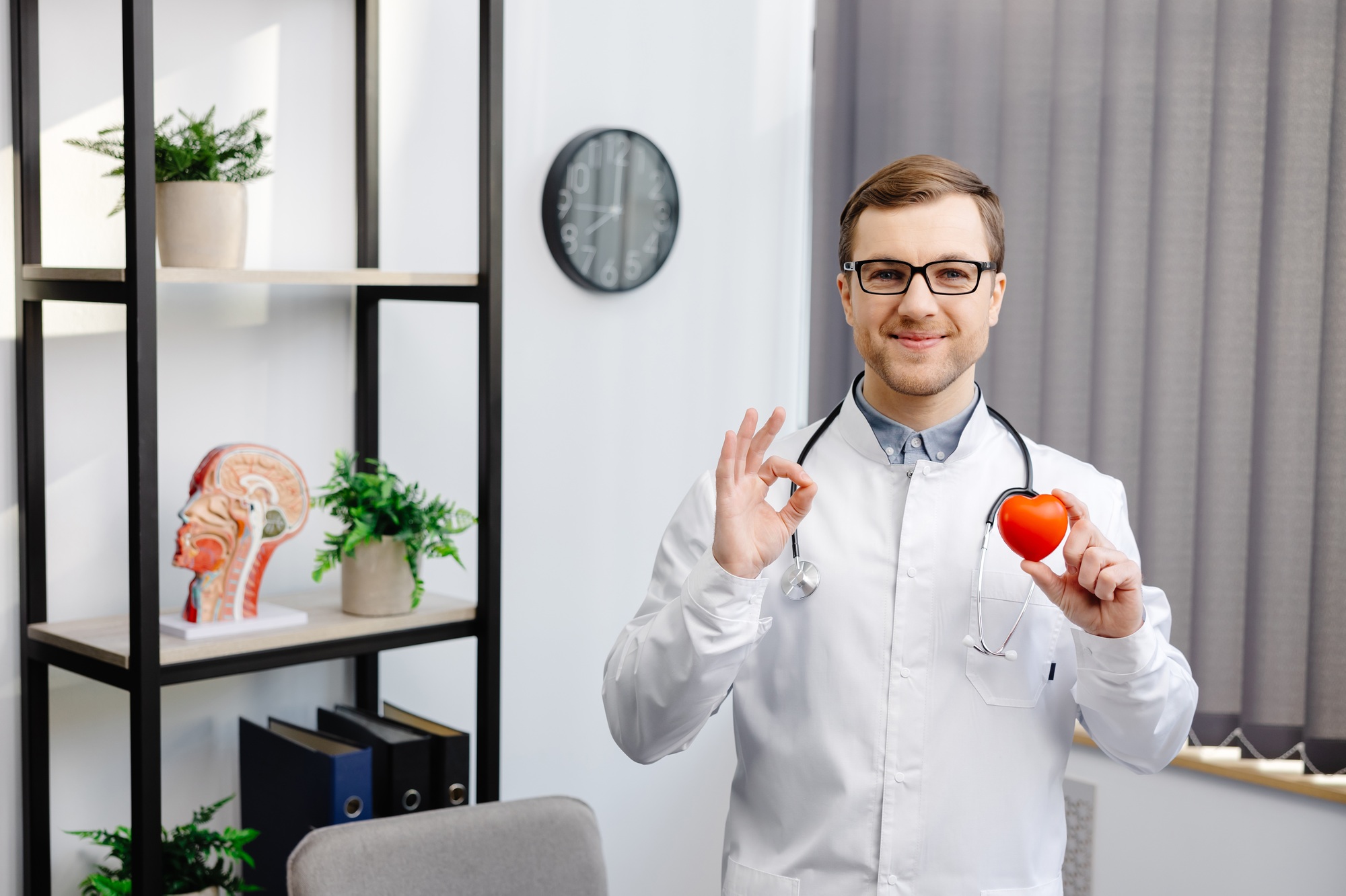 Doctor in white coat and glasses holding a red heart at hospital office